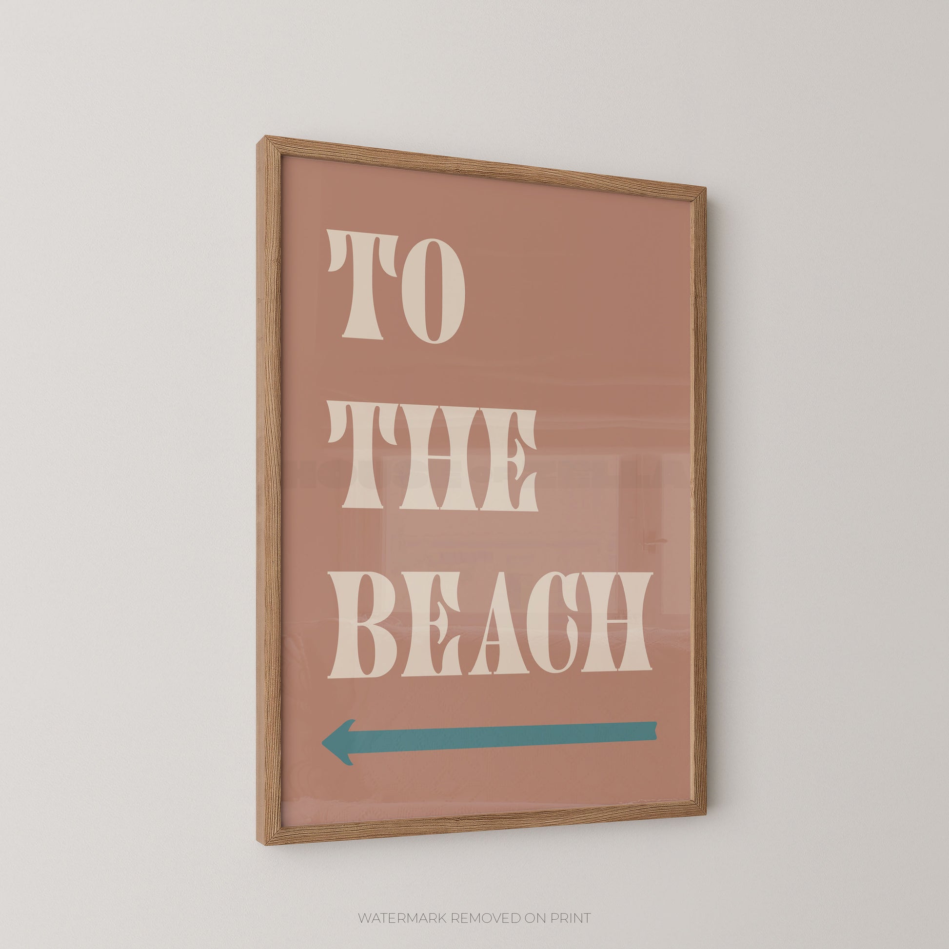 Framed wall art print with the words “TO THE BEACH” in large cream letters on a dusty pink background, plus a teal left-pointing arrow, shown hanging on a light neutral wall.