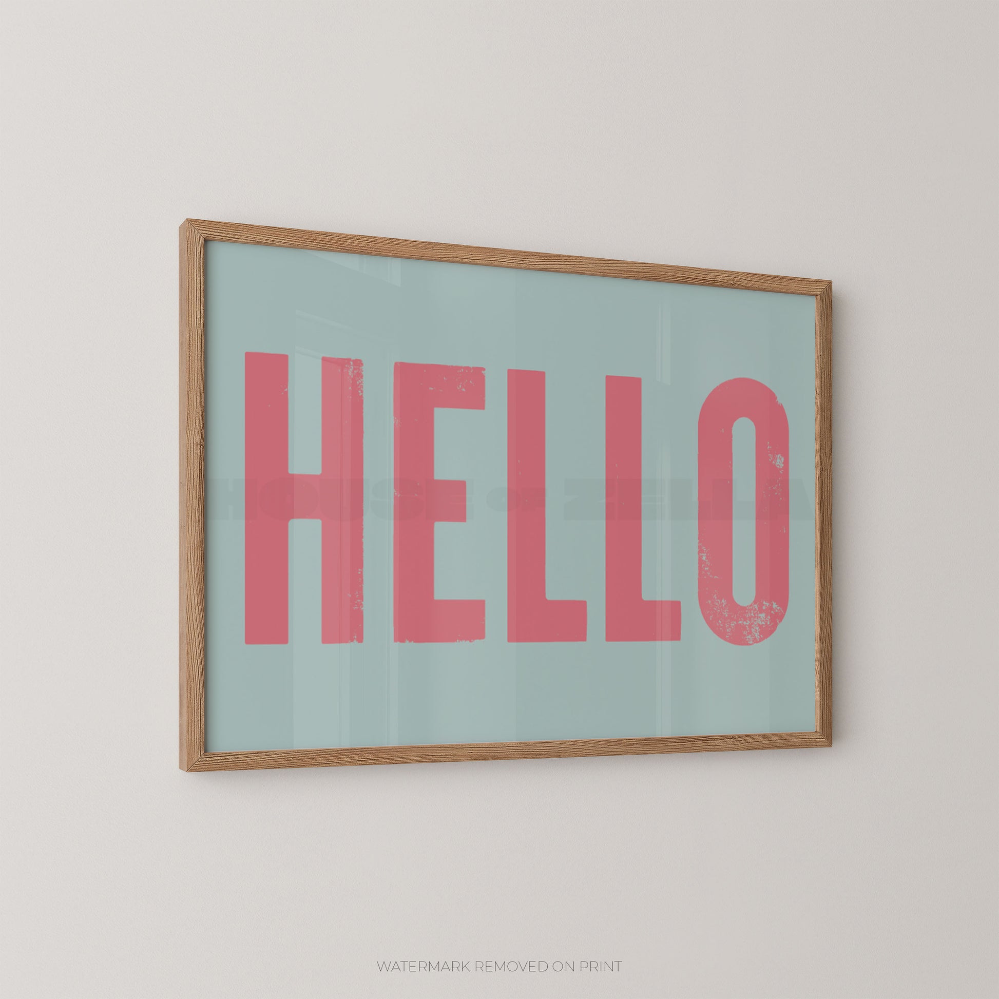 Framed typographic wall art hanging on a light wall, featuring the word "HELLO" in large pink letters on a pale blue background with a natural wood frame.