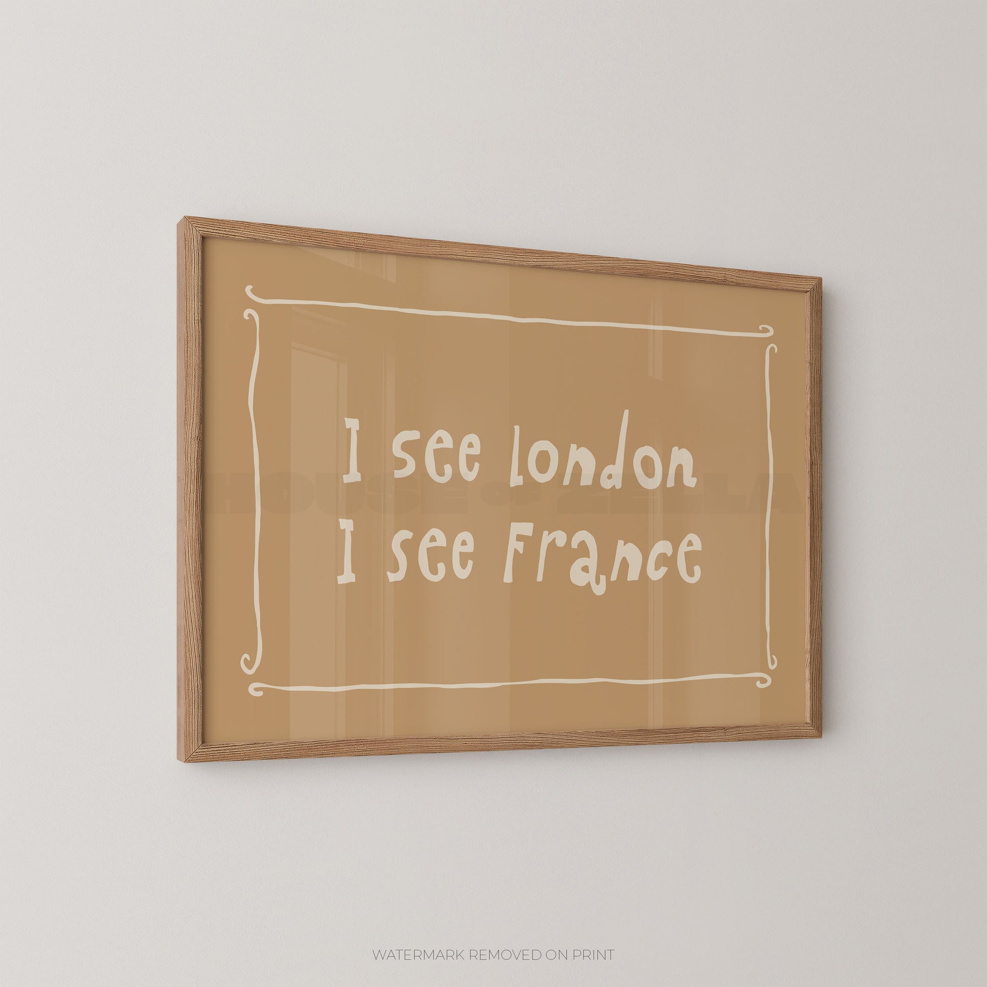 A framed typographic wall art print with the text 'I see london I see France' in white handwritten-style lettering on a tan background, enclosed in a light wood frame and mounted on a light gray wall.