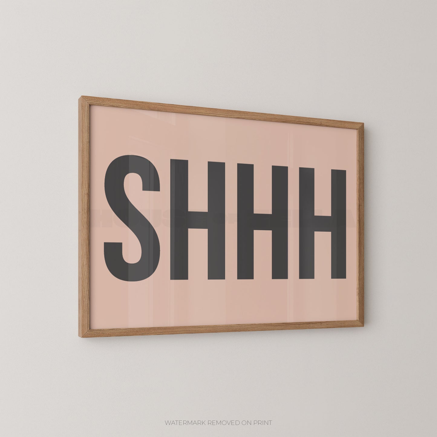 Framed typographic wall art on a light gray wall, featuring the word “SHHH” in large dark gray letters on a pale blush pink background with a natural wood frame.