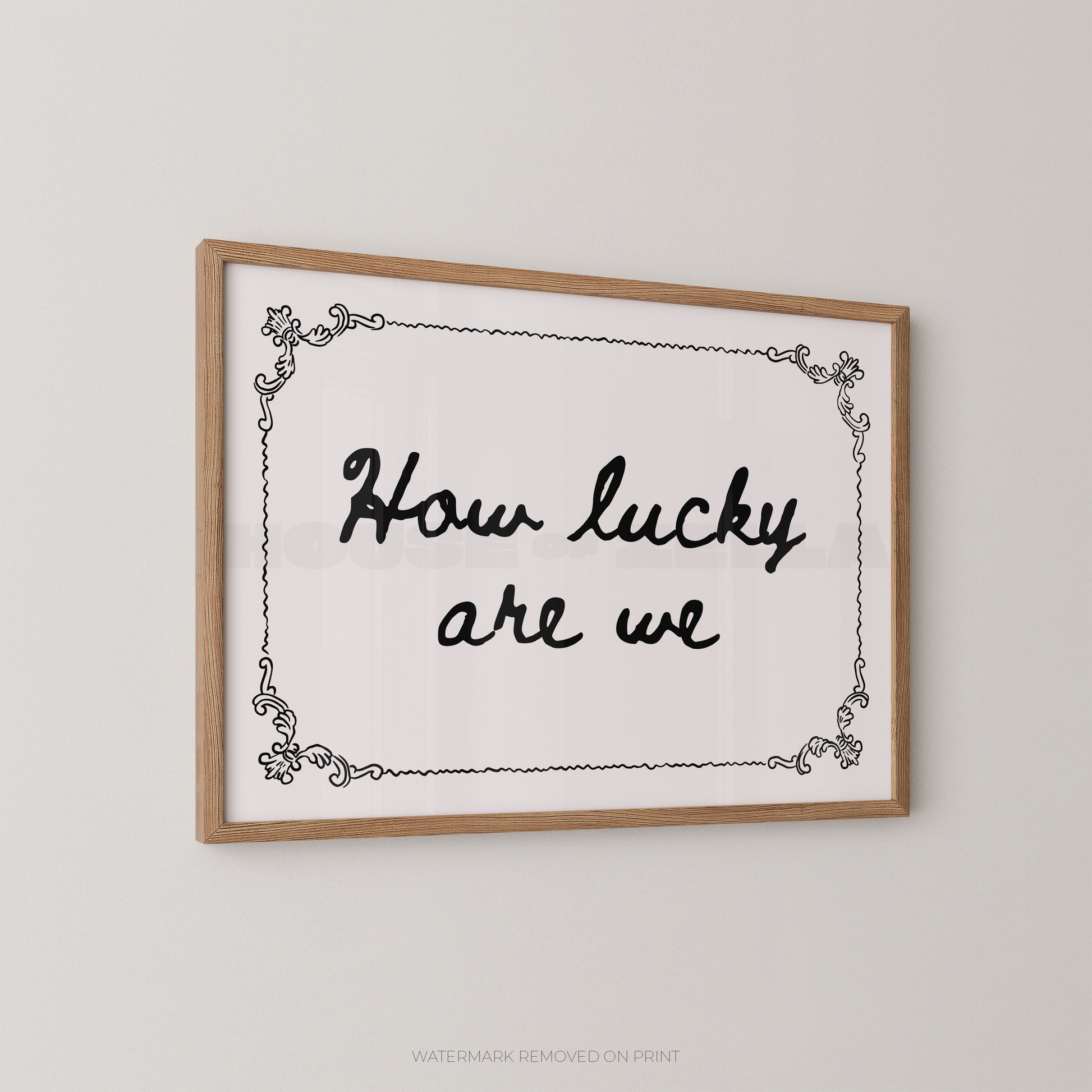 Framed wall art print with the phrase “How lucky are we” in black script inside a light wood frame, shown hanging on a plain light gray wall.