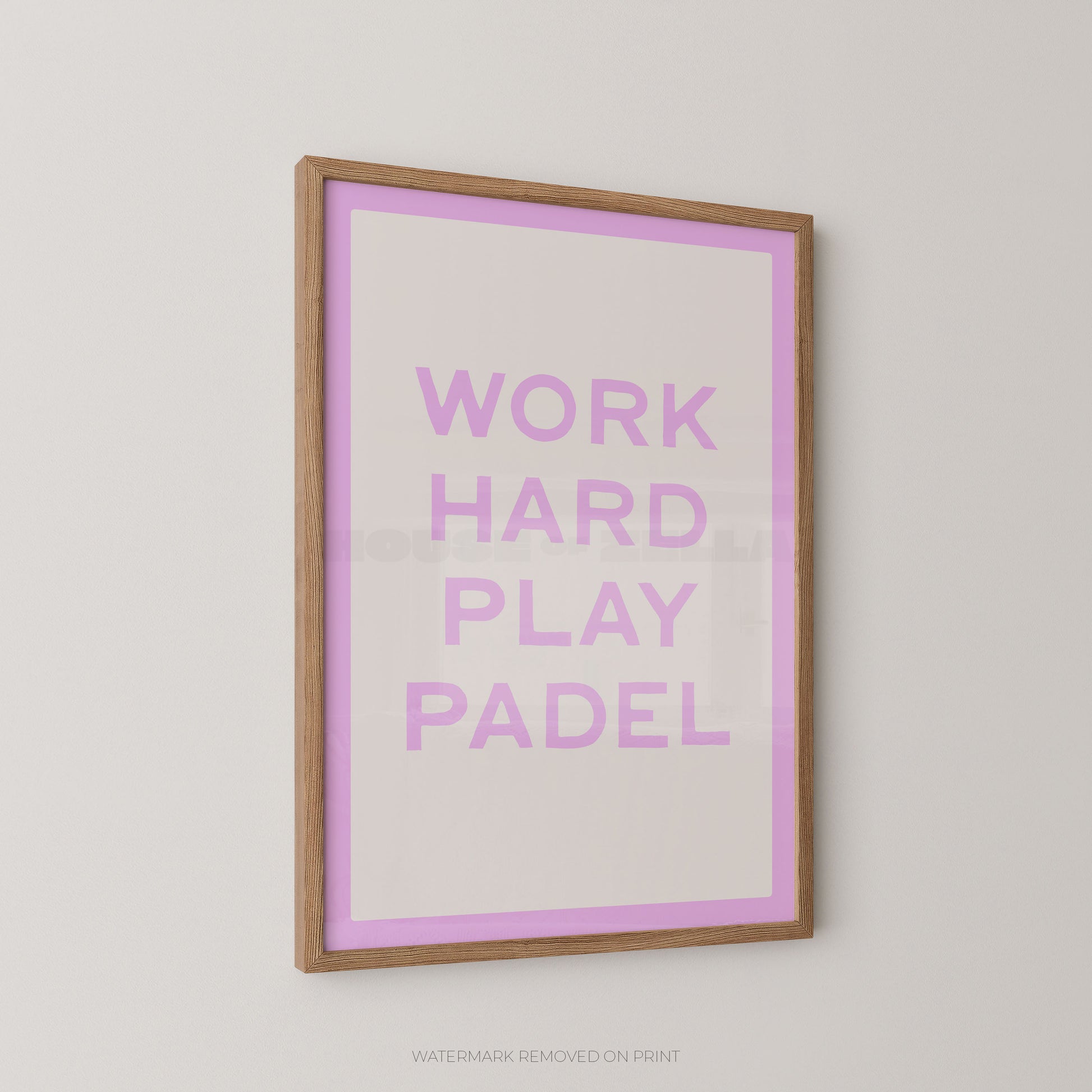 A framed typographic wall art print with the words “WORK HARD PLAY PADEL” in lavender-purple lettering on a white background with a lavender border, set in a light natural wood frame against a light gray wall.
