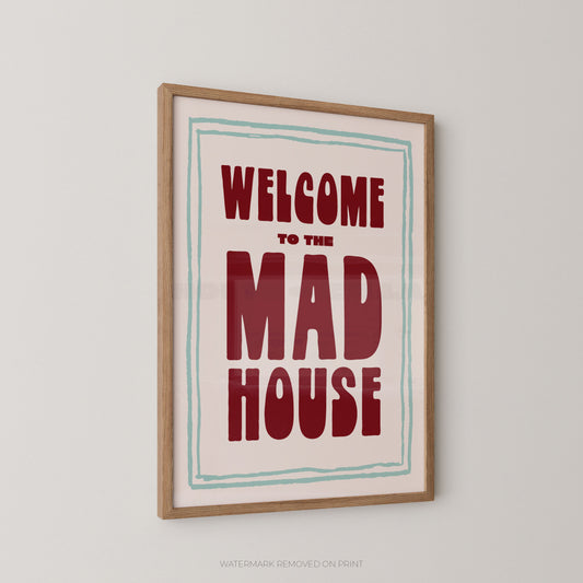 Framed wall art print with the text 'WELCOME TO THE MAD HOUSE' in bold red lettering on a cream background,  with a pale blue borderenclosed in a light wood frame and shown hanging on a light gray wall.