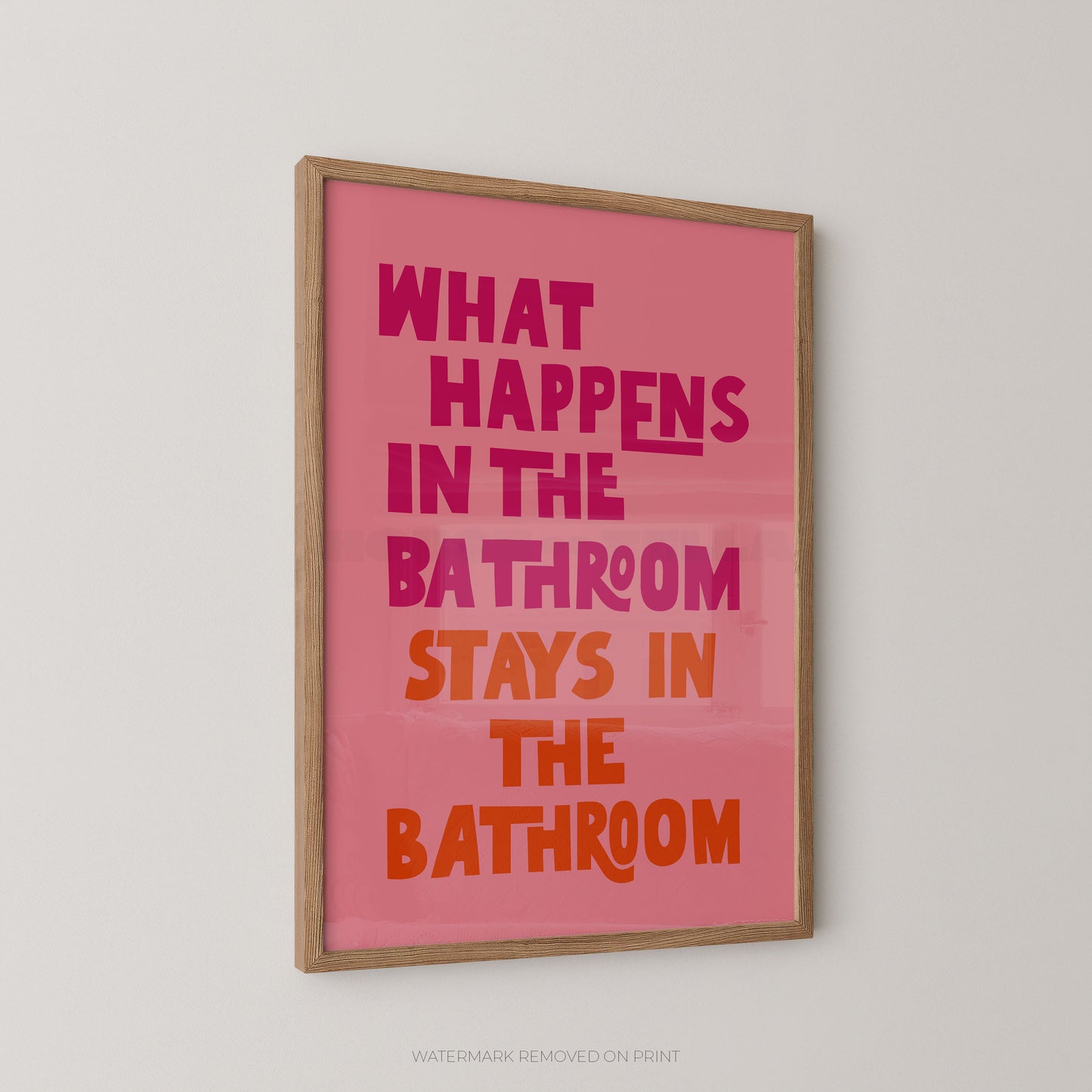 Funny bathroom wall art print in a wood frame – playful chunky pink and orange typography on a pink background reading "What Happens In The Bathroom Stays In The Bathroom"