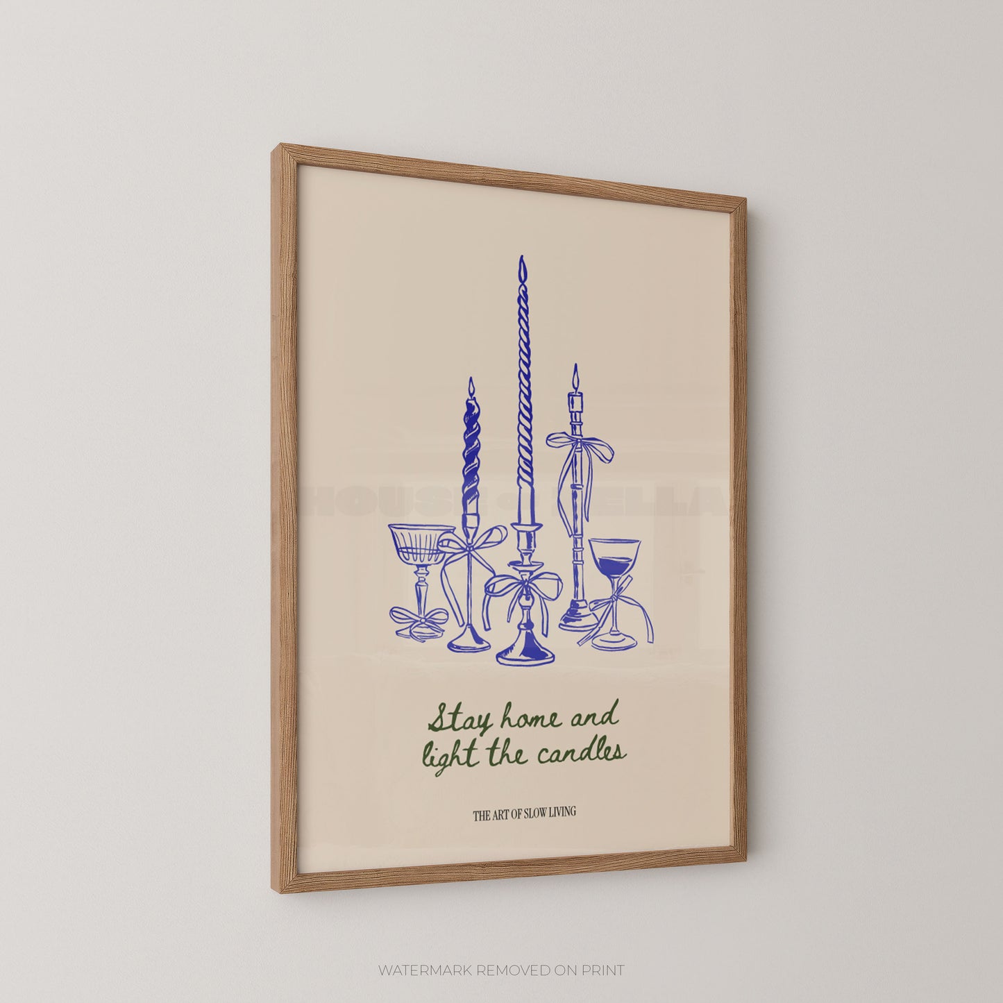Framed wall art print featuring a blue line drawing of candles and goblets with the text 'Stay home and light the candles' and 'THE ART OF SLOW LIVING' on a warm cream background, shown hanging on a light wall.