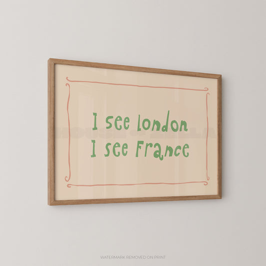 Framed typographic wall art hanging on a light gray wall, featuring the handwritten-style text 'I see London I see France' in green inside a coral-pink decorative border on a warm cream background, with a natural wood frame.