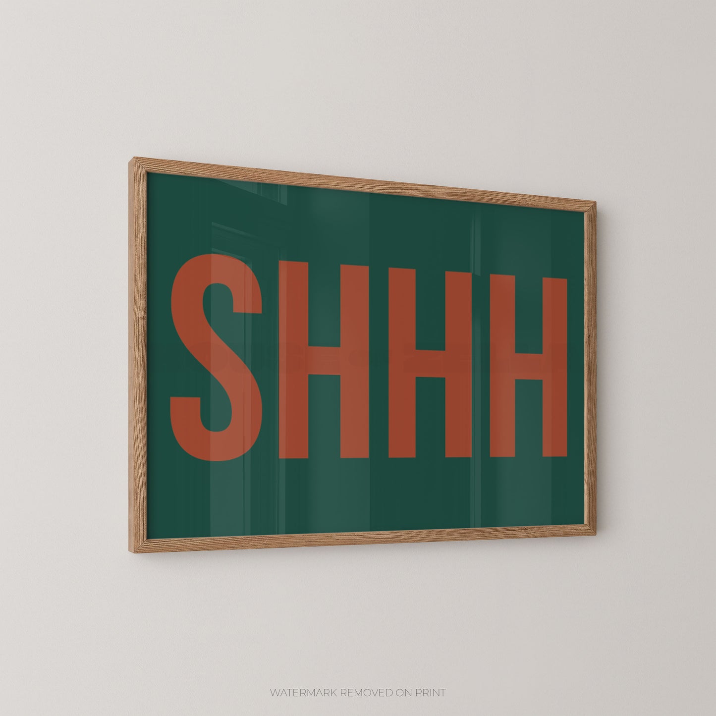Framed wall art print with the word 'SHHH' in large orange-red letters on a dark green background, shown hanging on a light gray wall.