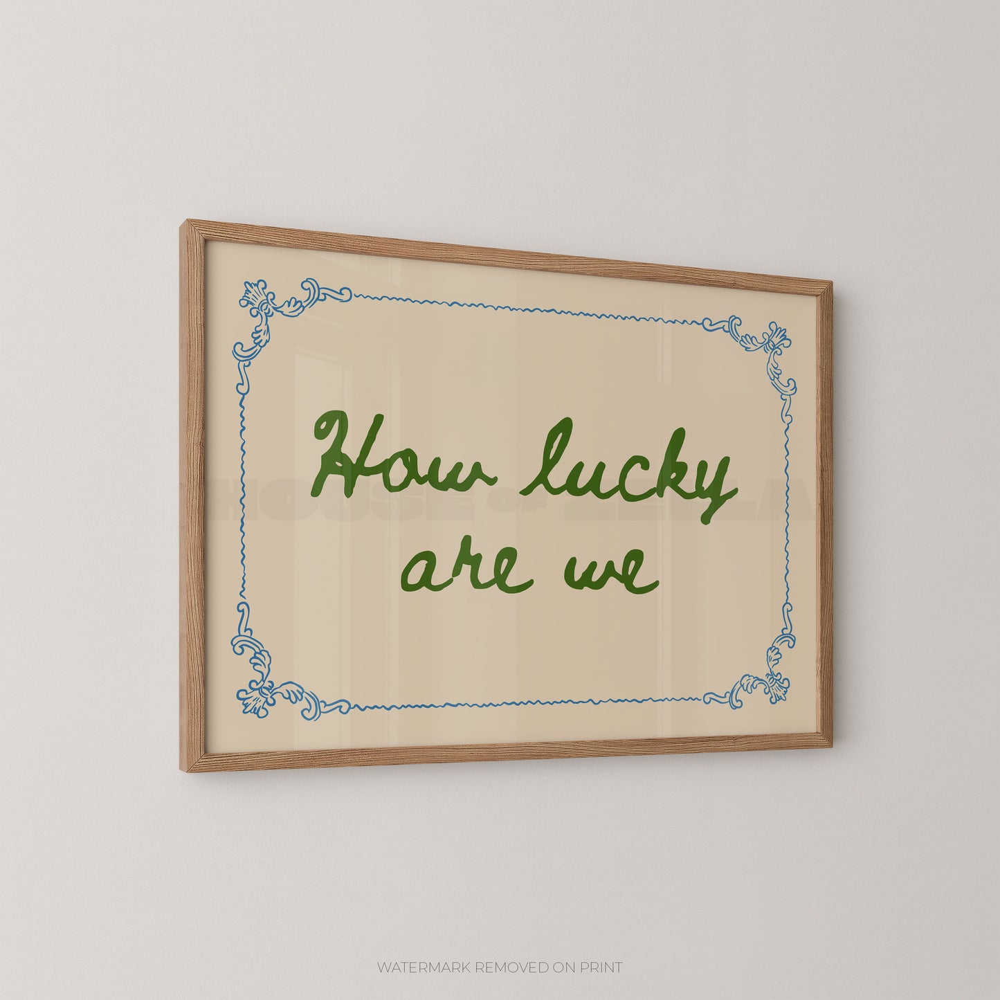Framed wall art with the phrase “How lucky are we” in green script, set on a cream background with a light blue decorative border and a natural wood frame, shown hanging on a light gray wall.