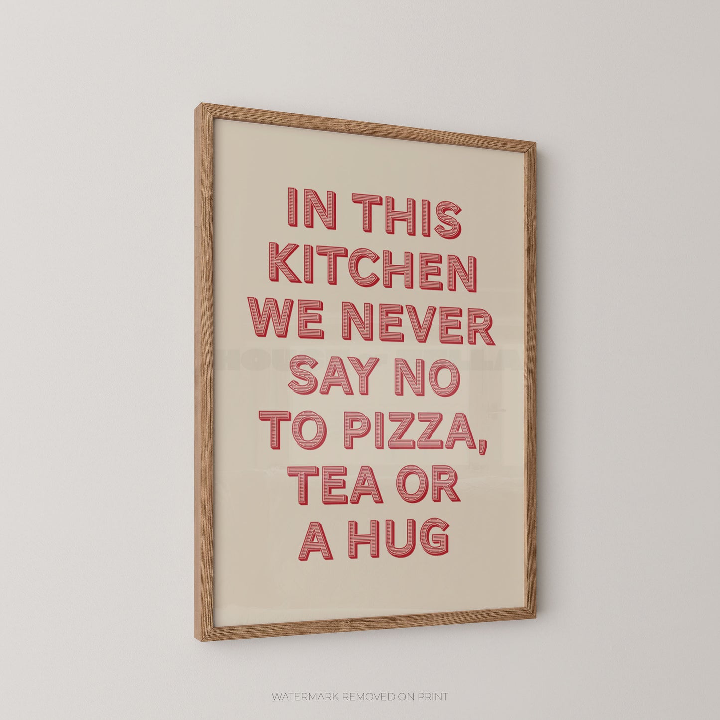 A framed typographic wall art print hanging on a light gray wall. The print has a cream background with raised coral-red text reading, "IN THIS KITCHEN WE NEVER SAY NO TO PIZZA, TEA OR A HUG," in a light natural wood frame.