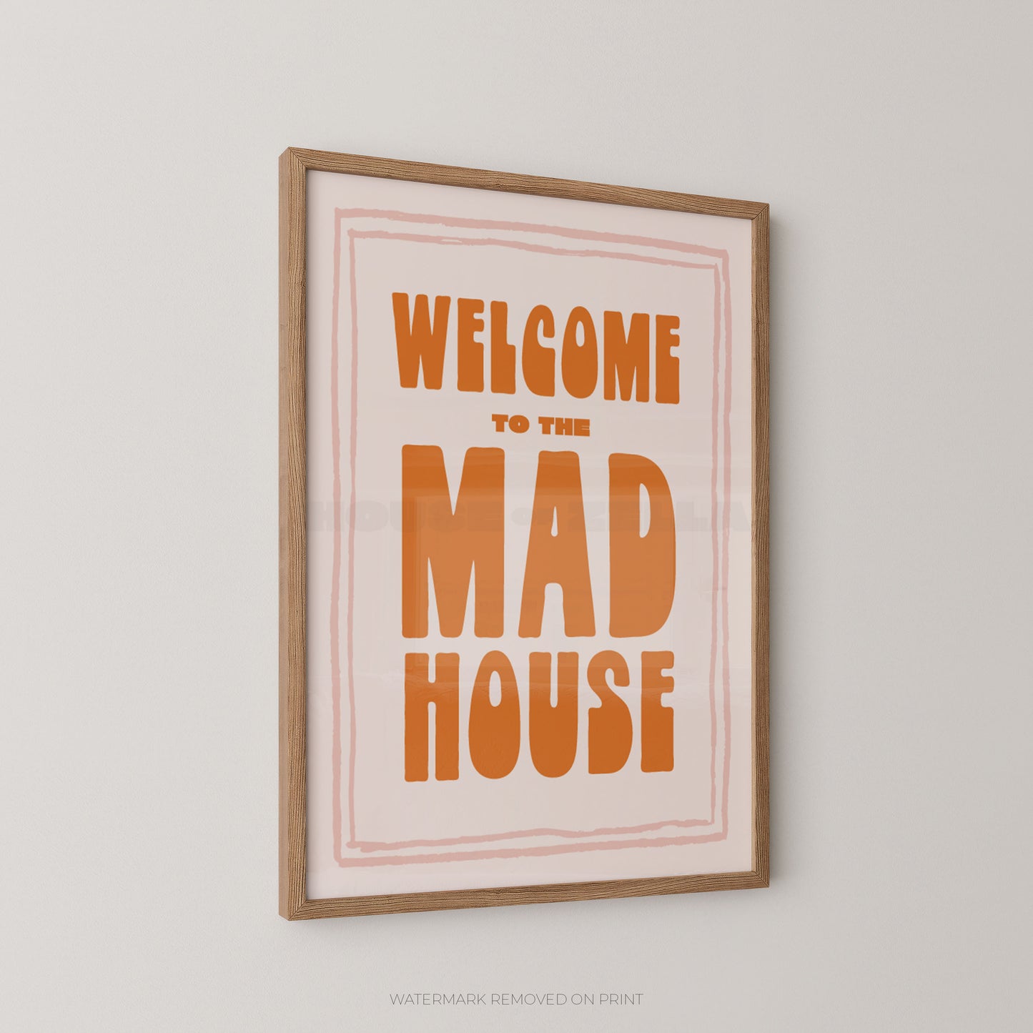 A framed typographic wall art print reading “WELCOME TO THE MAD HOUSE” in orange lettering on a pale pink background, with a light wood frame, shown hanging against a light gray wall.