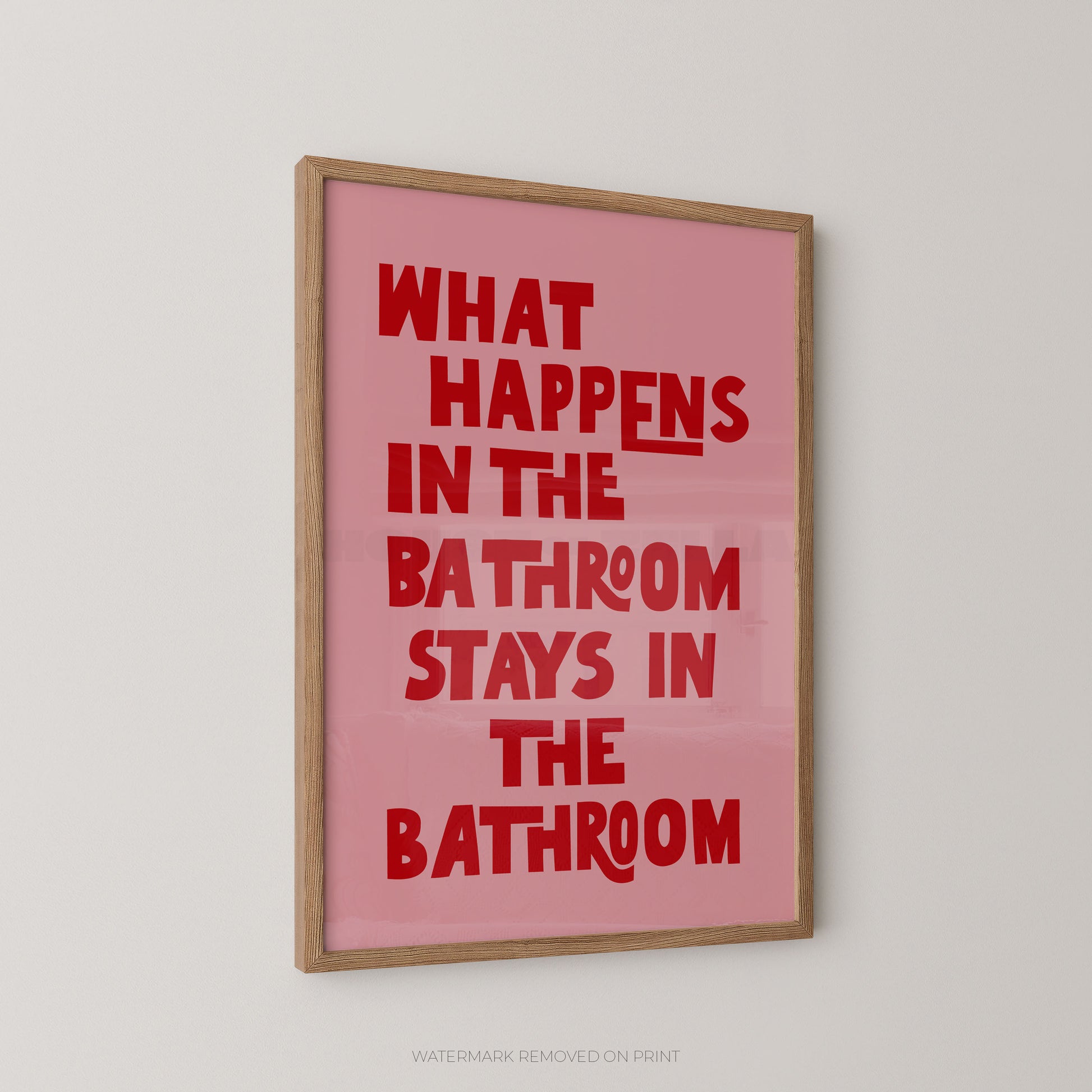 Funny bathroom wall art print in a wood frame – bold pink and red chunky typography on a light pink background reading "What Happens In The Bathroom Stays In The Bathroom"