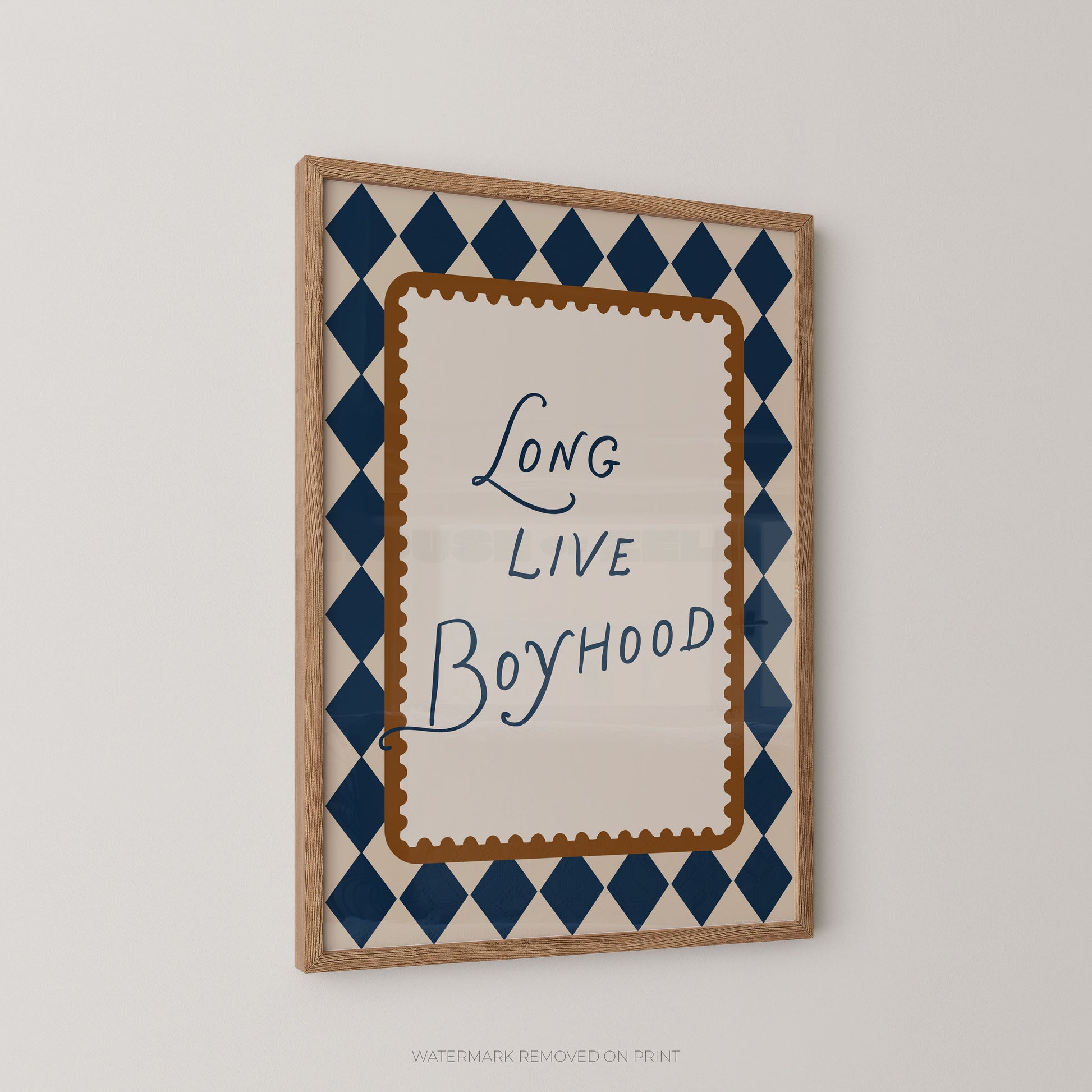 Framed wall art print with the phrase 'Long Live Boyhood' in blue script inside a cream central panel, surrounded by a brown scalloped border and a navy-and-cream harlequin diamond pattern, shown hanging on a light gray wall.