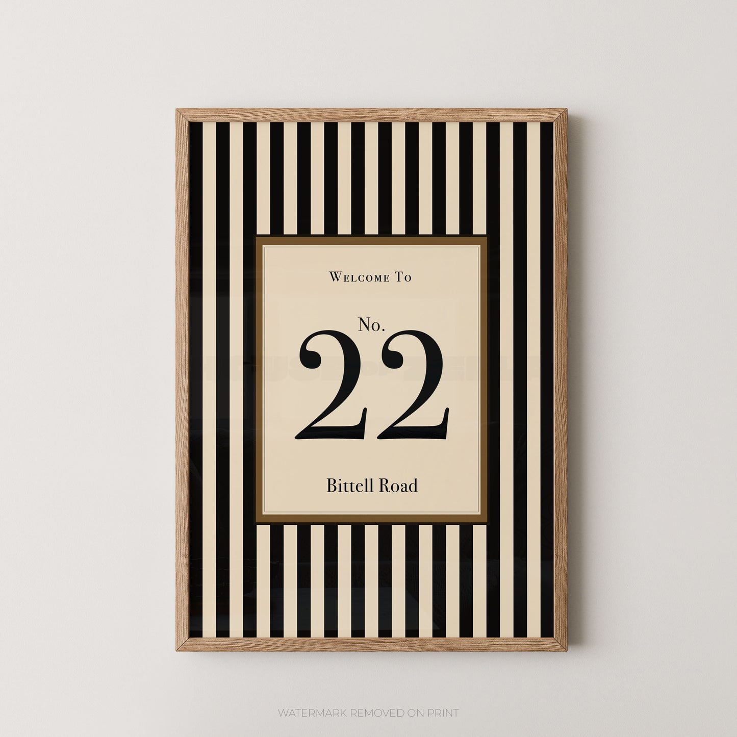 Framed wall art print with a black and cream vertical stripe pattern, featuring a central cream panel with the text 'WELCOME TO No. 22 Bitell Road' in black serif lettering, shown against a light gray wall.