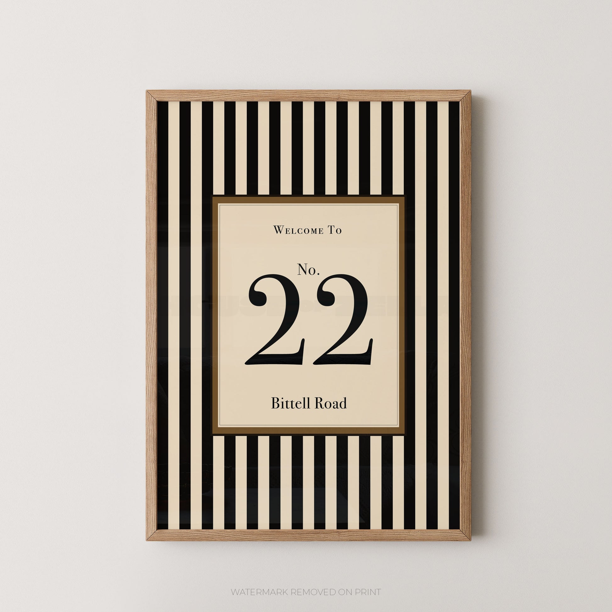 Framed wall art print with a black and cream vertical stripe pattern, featuring a central cream panel with the text 'WELCOME TO No. 22 Bitell Road' in black serif lettering, shown against a light gray wall.