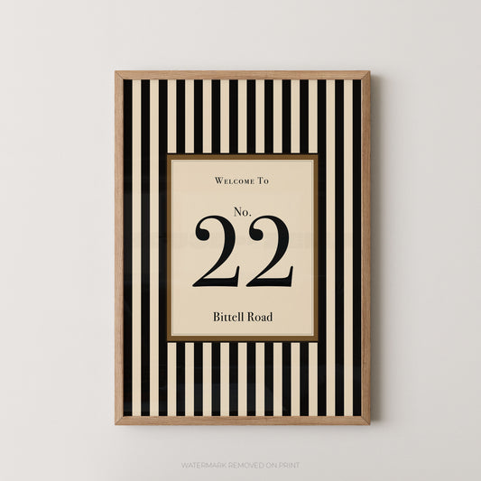 Framed wall art print with a black and cream vertical stripe pattern, featuring a central cream panel with the text 'WELCOME TO No. 22 Bitell Road' in black serif lettering, shown against a light gray wall.