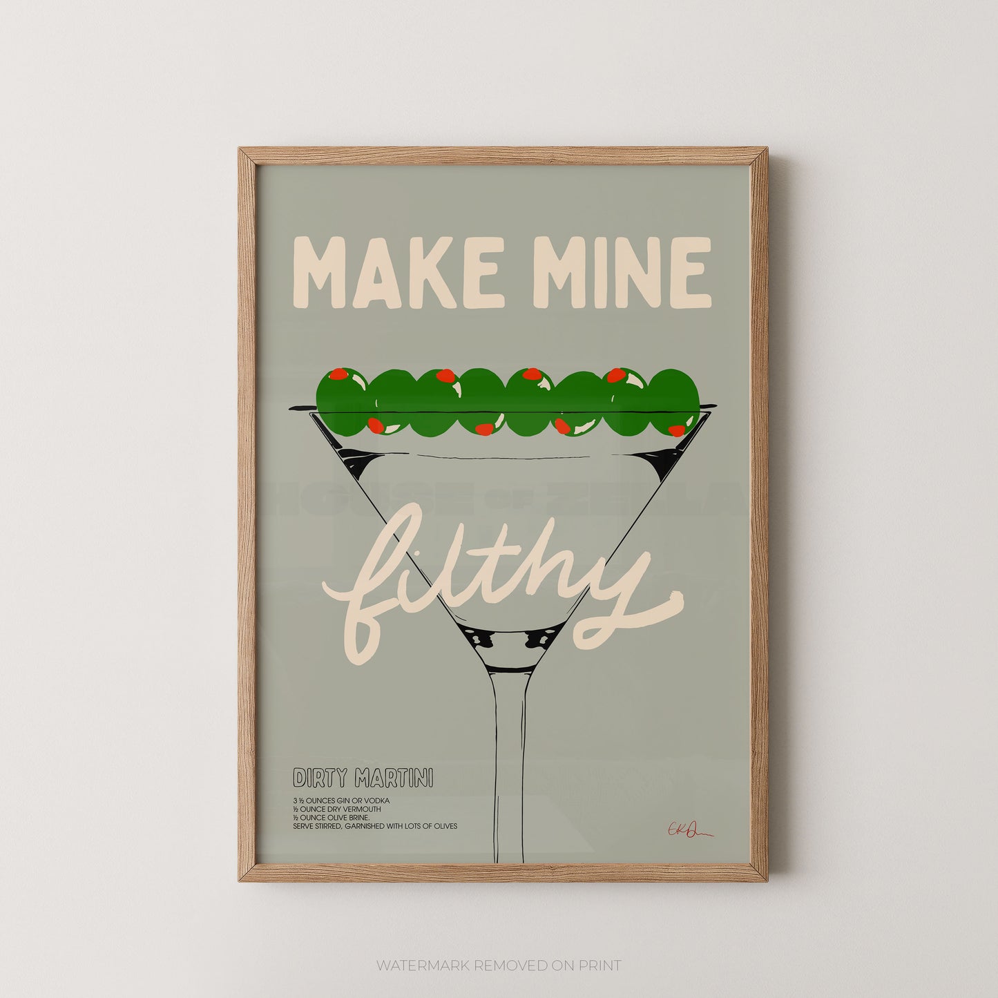 A framed art print featuring a stylized martini glass with olives and the text 'MAKE MINE filthy' on a muted sage background, shown against a light neutral wall.
