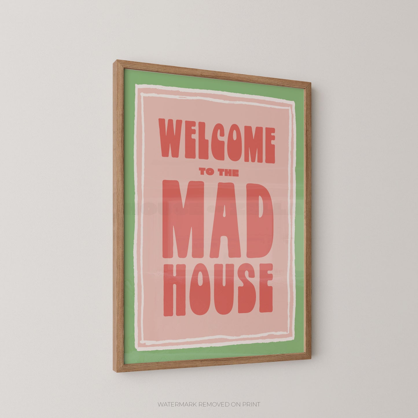 A framed art print featuring the text 'WELCOME TO THE MAD HOUSE' in coral-red lettering on a pink background, bordered by a green inner frame and a natural wood outer frame, hanging on a light gray wall.