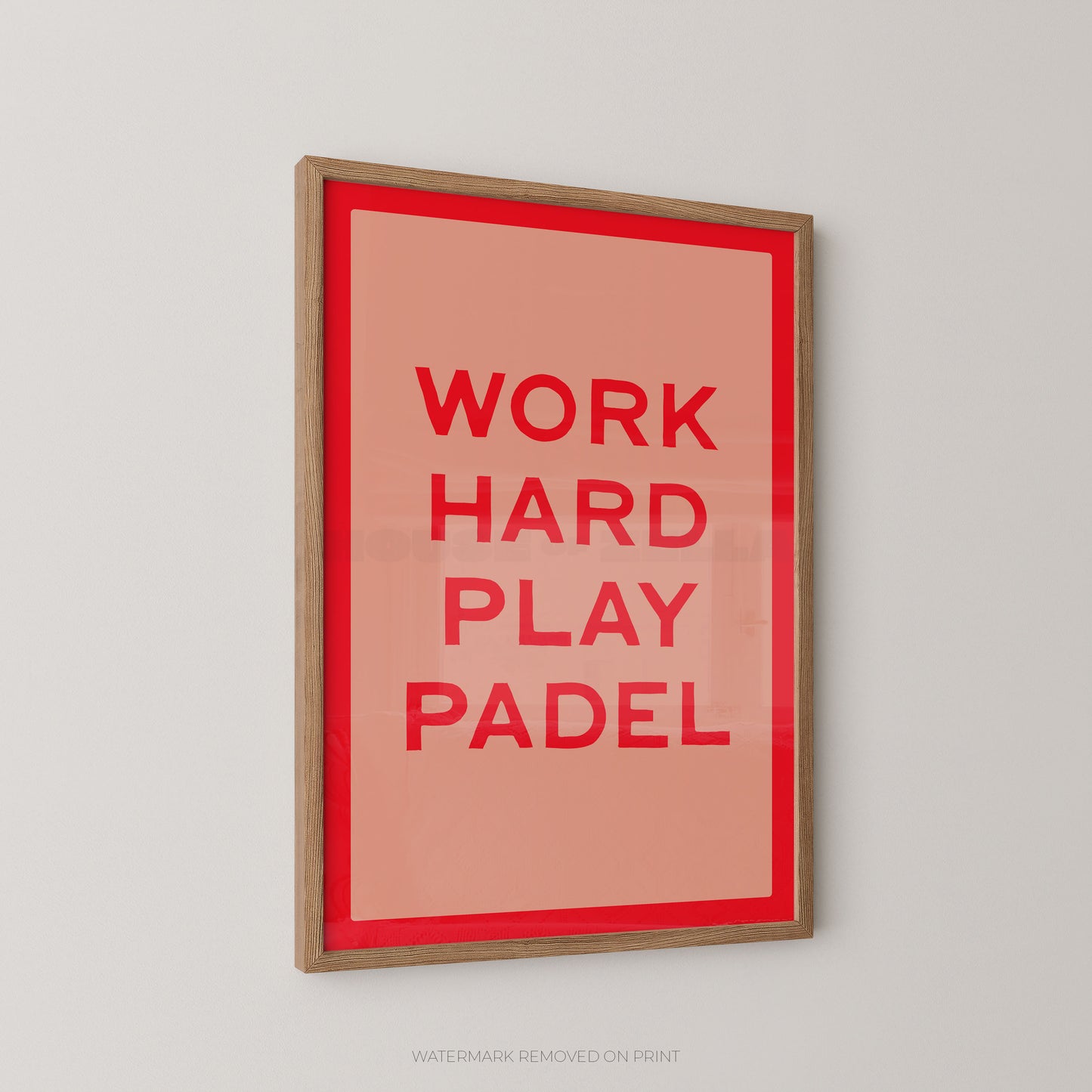 A framed typographic wall art print with the words 'WORK HARD PLAY PADEL' in red on a coral background, surrounded by a red inner border and a light wood frame, shown hanging on a light gray wall.