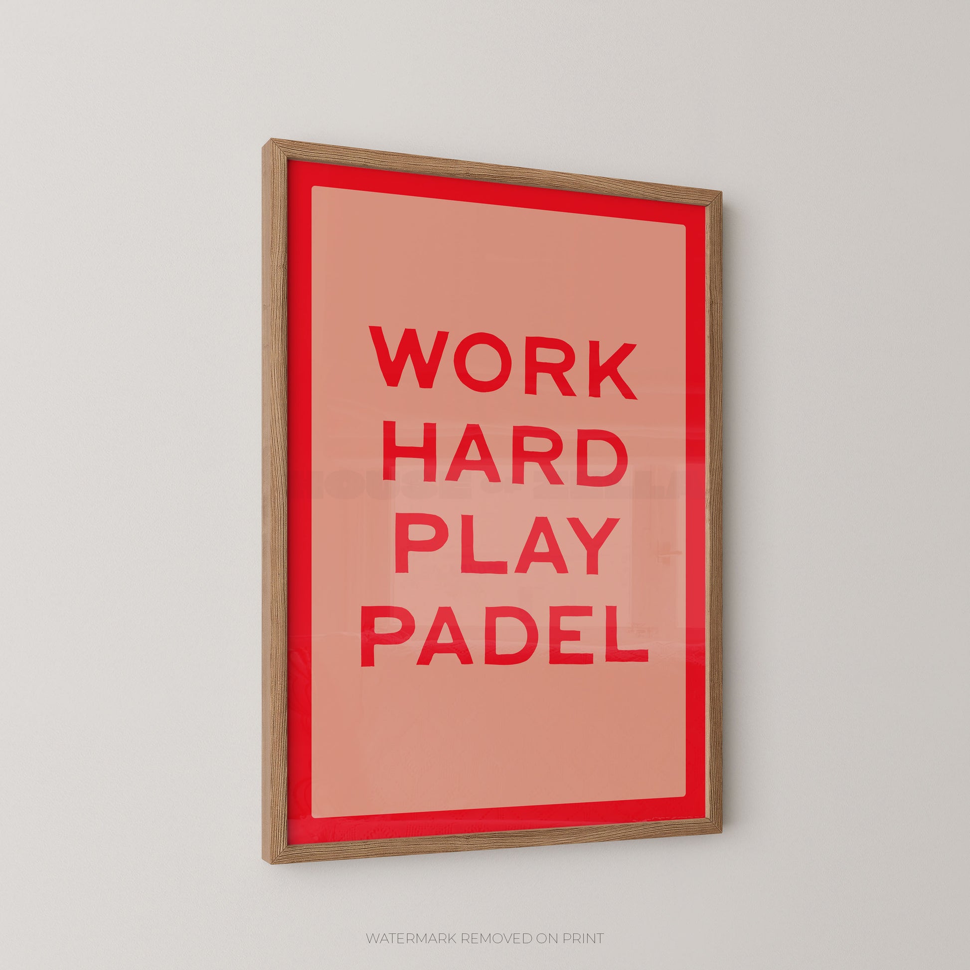 A framed typographic wall art print with the words 'WORK HARD PLAY PADEL' in red on a coral background, surrounded by a red inner border and a light wood frame, shown hanging on a light gray wall.
