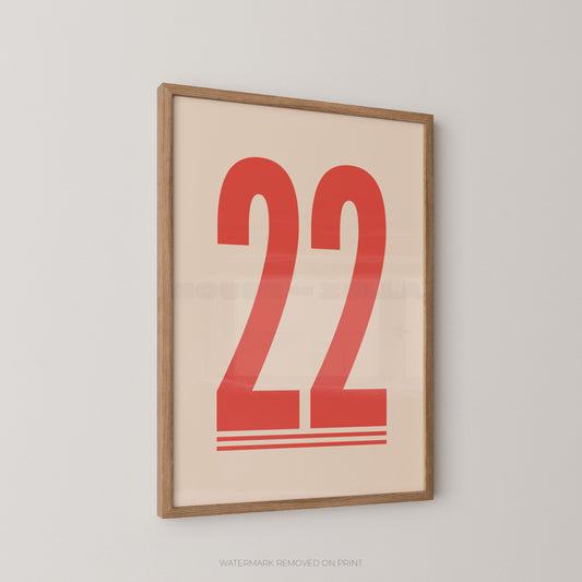 Framed minimalist wall art print featuring the red number 22 on a warm cream background, enclosed in a light natural wood frame and shown hanging on a light gray wall.