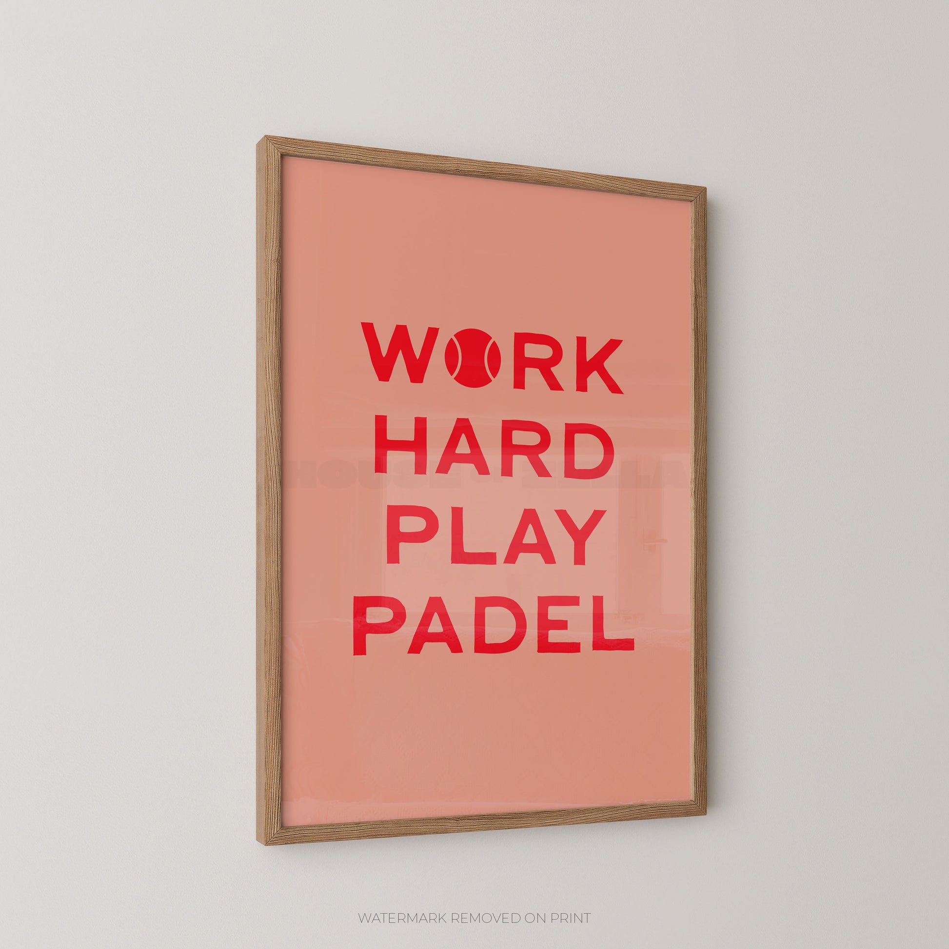 A framed wall art print with a peach background and bold red text reading “WORK HARD PLAY PADEL,” with the O replaced by a tennis ball icon. The print is mounted in a light natural wood frame and shown hanging on a light gray wall.
