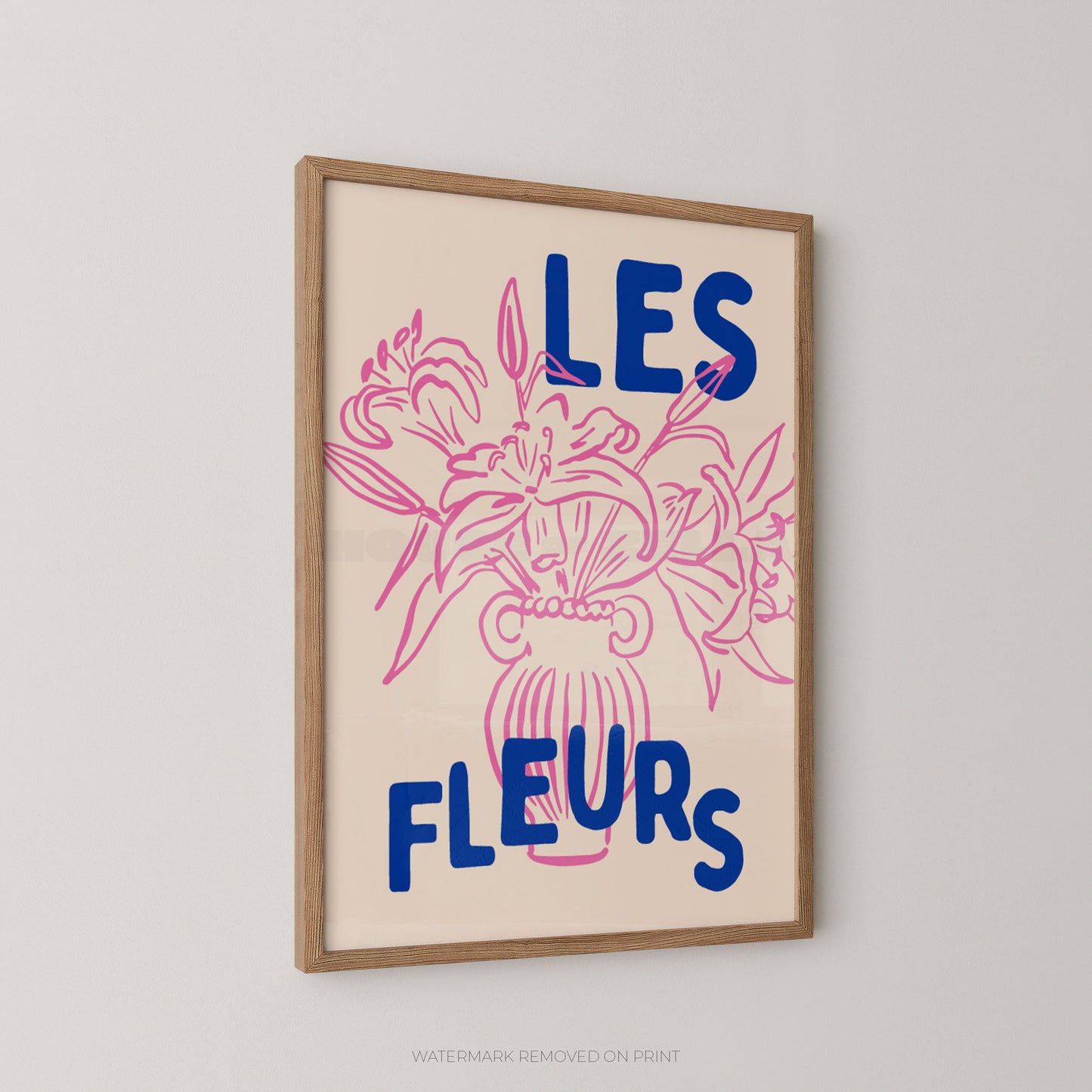 A framed wall art print with the French text “LES FLEURS” in blue over a pink floral illustration on a cream background, shown hanging on a light gray wall.