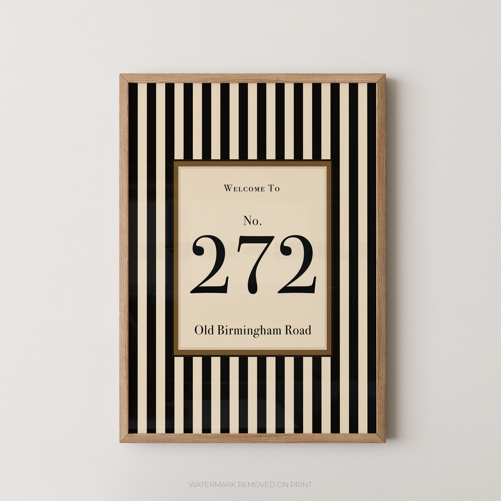 A framed vertical wall art print with black and cream stripes, featuring a central cream panel with the text 'Welcome To No. 272 Old Birmingham Road' and a light wood frame, shown against a light gray wall.