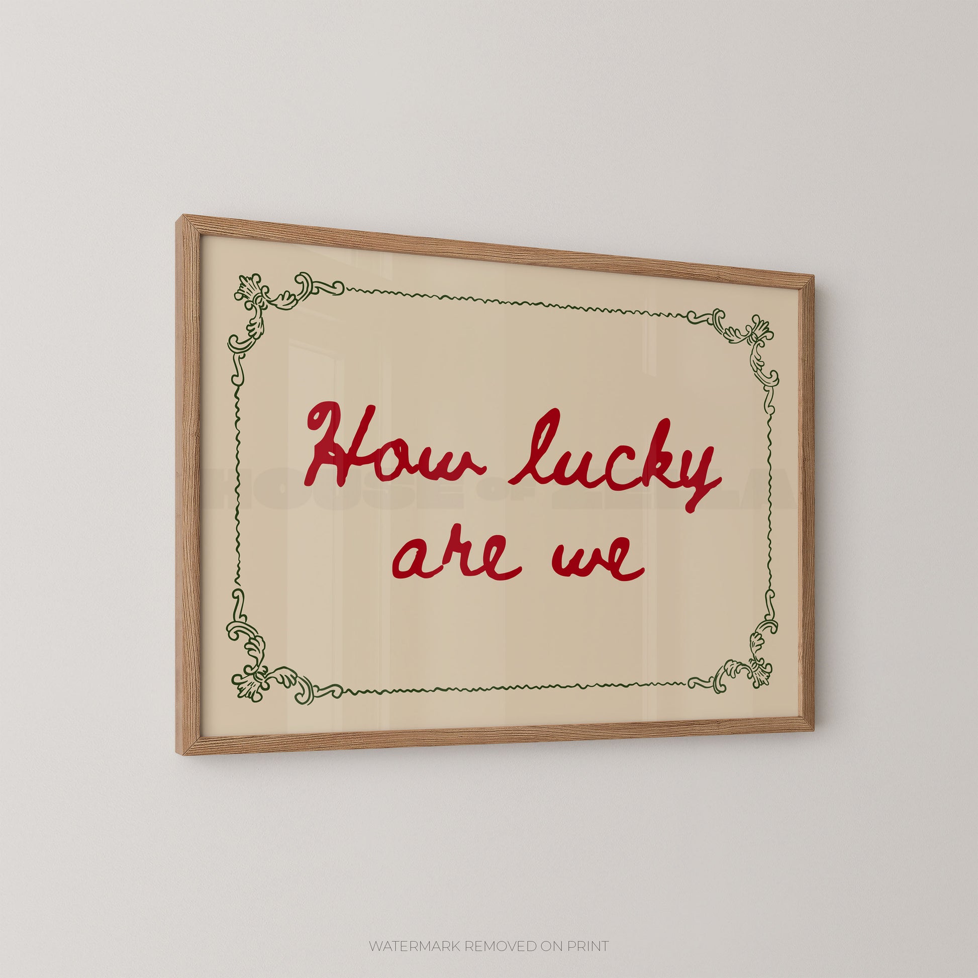 A framed typographic wall art print with the phrase “How lucky are we” in red script, set within a light wood frame and a pale cream background, shown hanging on a light gray wall.