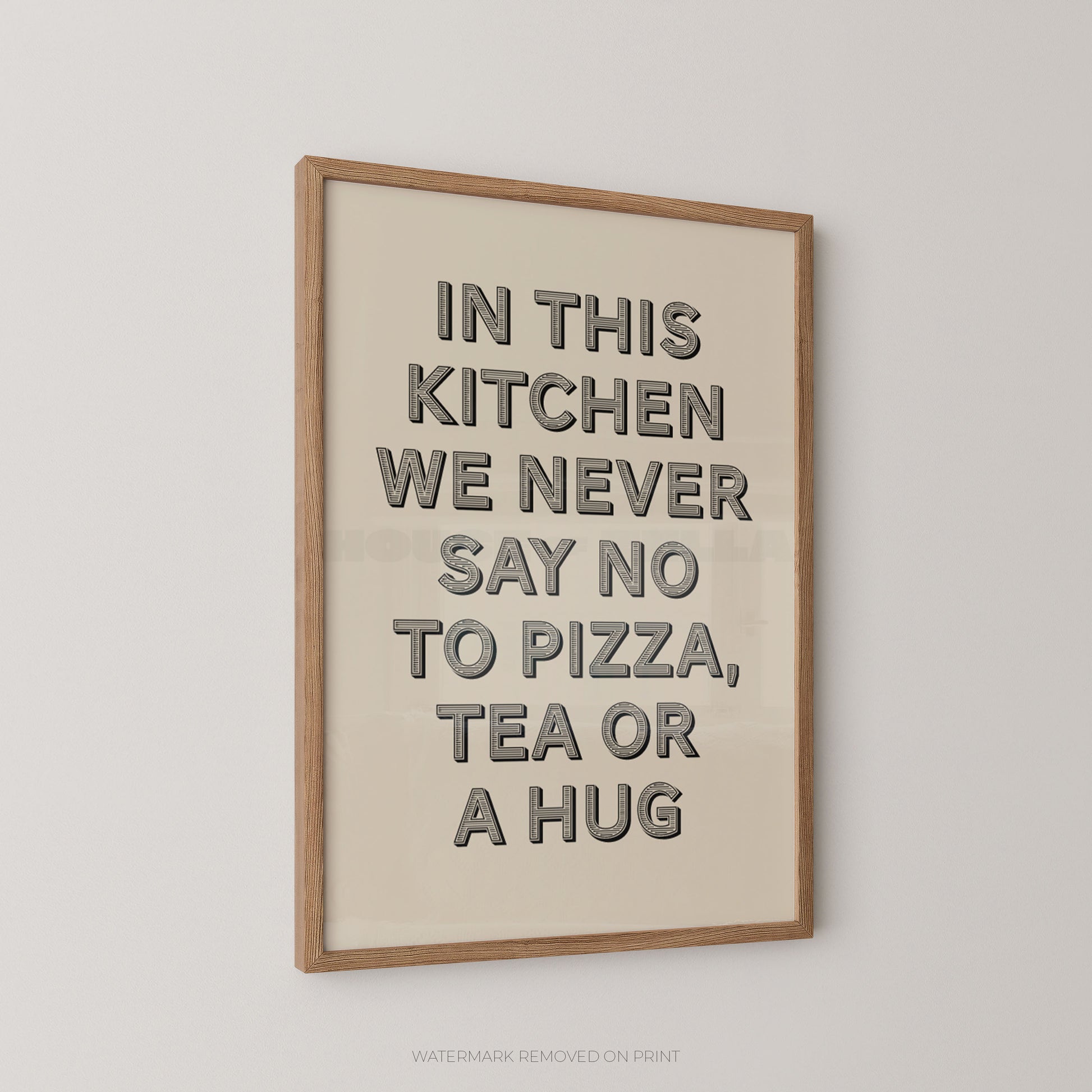 A framed typographic wall art print with a cream background and black raised lettering that reads, 'IN THIS KITCHEN WE NEVER SAY NO TO PIZZA, TEA OR A HUG,' shown hanging on a light neutral wall.