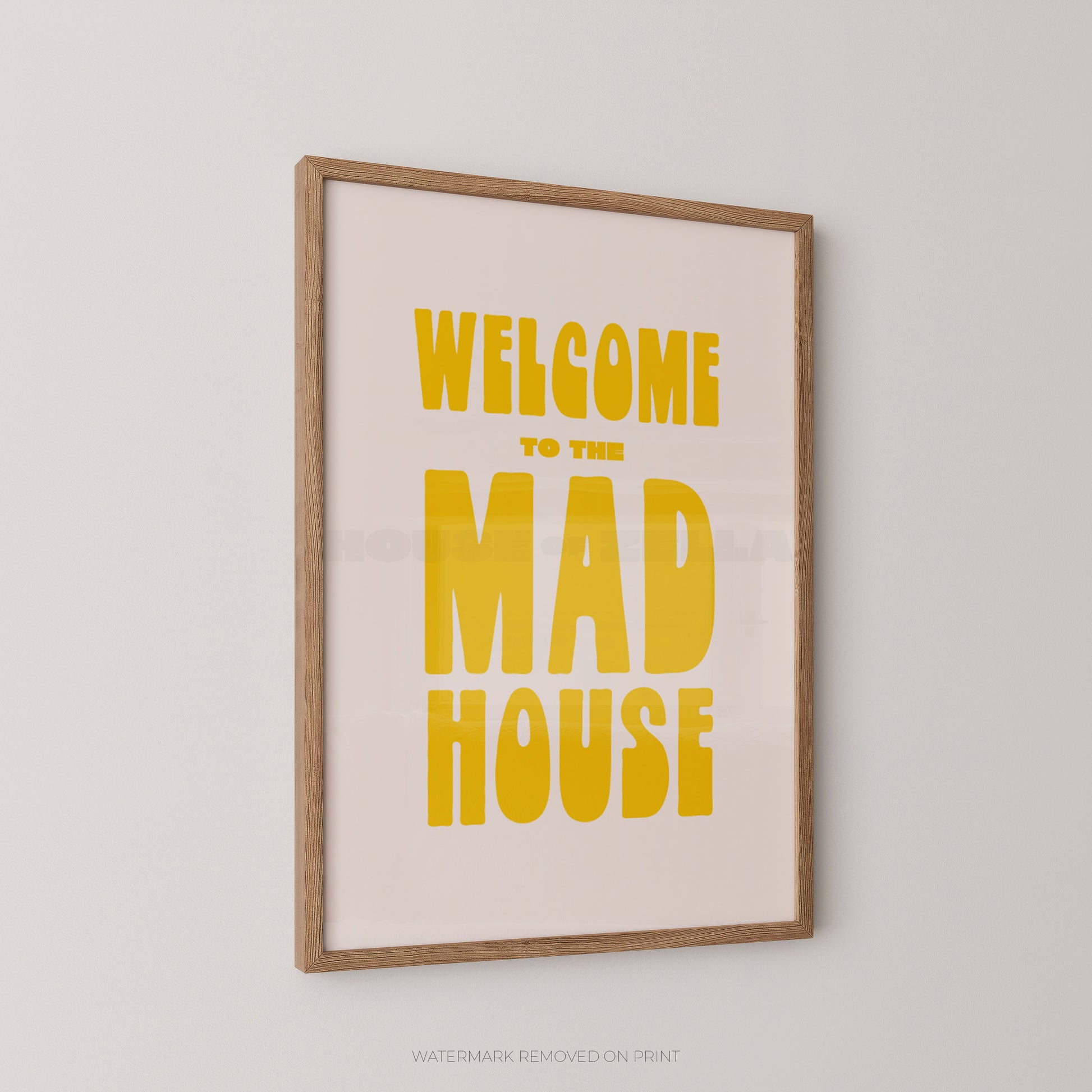 A framed typographic wall art print reading “WELCOME TO THE MAD HOUSE” in bold yellow text on a pale pink background, shown hanging on a light gray wall.