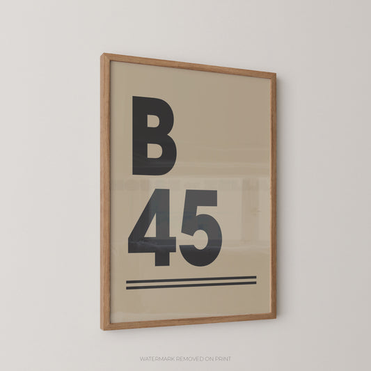 Framed minimalist typographic wall art hanging on a light gray wall. The print features a large black letter B above the number 45, with thin horizontal lines beneath, on a warm beige background.