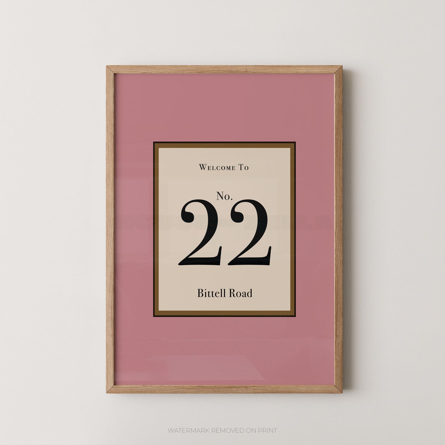 Framed wall art print with a dusty pink mat and a light wood frame, featuring a cream central panel with the text 'WELCOME TO No. 22 Bittell Road' in black serif lettering.