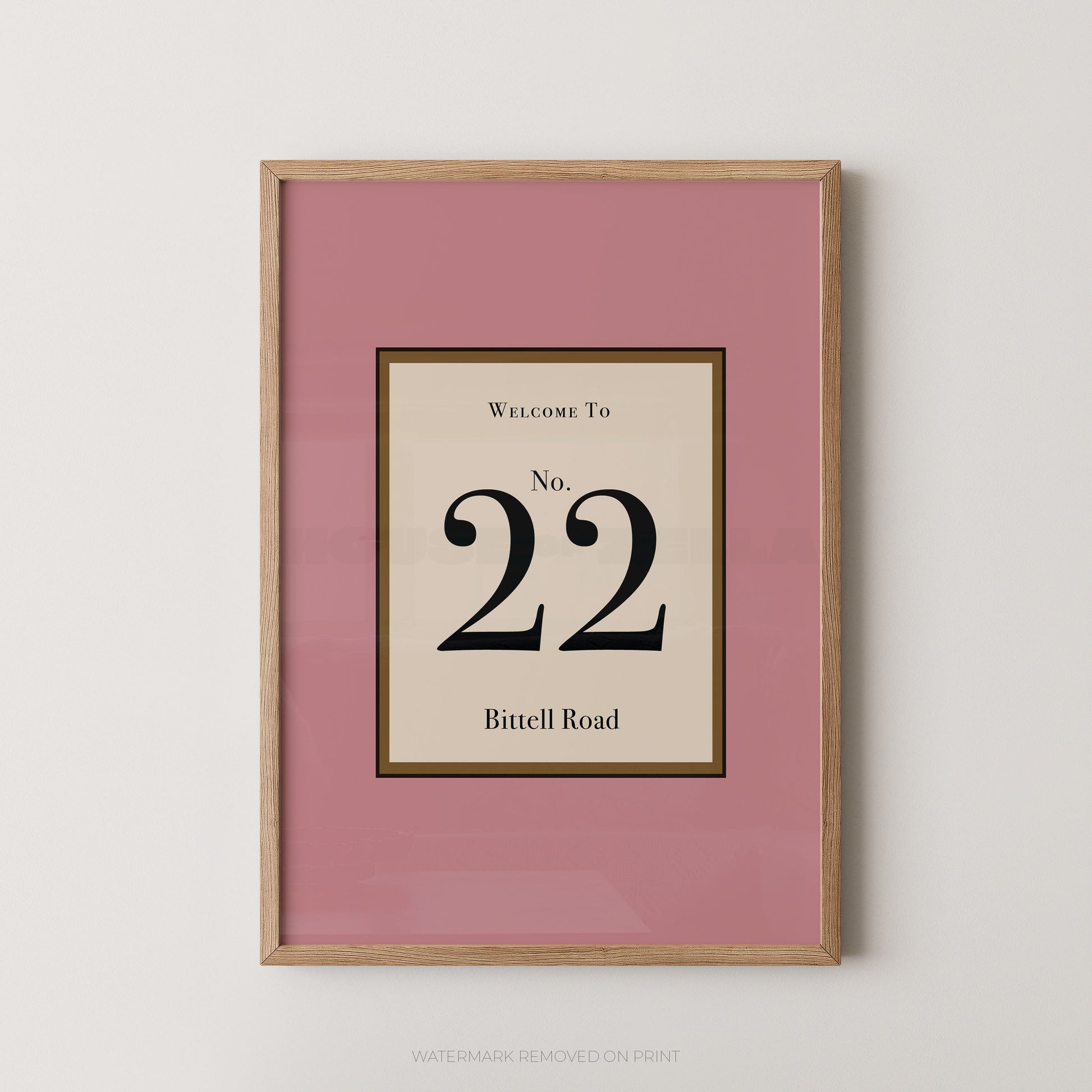Framed wall art print with a dusty pink mat and a light wood frame, featuring a cream central panel with the text 'WELCOME TO No. 22 Bittell Road' in black serif lettering.