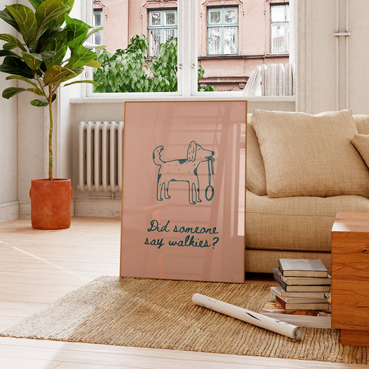 A vertical framed art print leaning against a beige sofa in a bright living room. The print has a dusty pink background with a teal line drawing of a dog holding a leash and the text 'Did someone say walkers?'.
