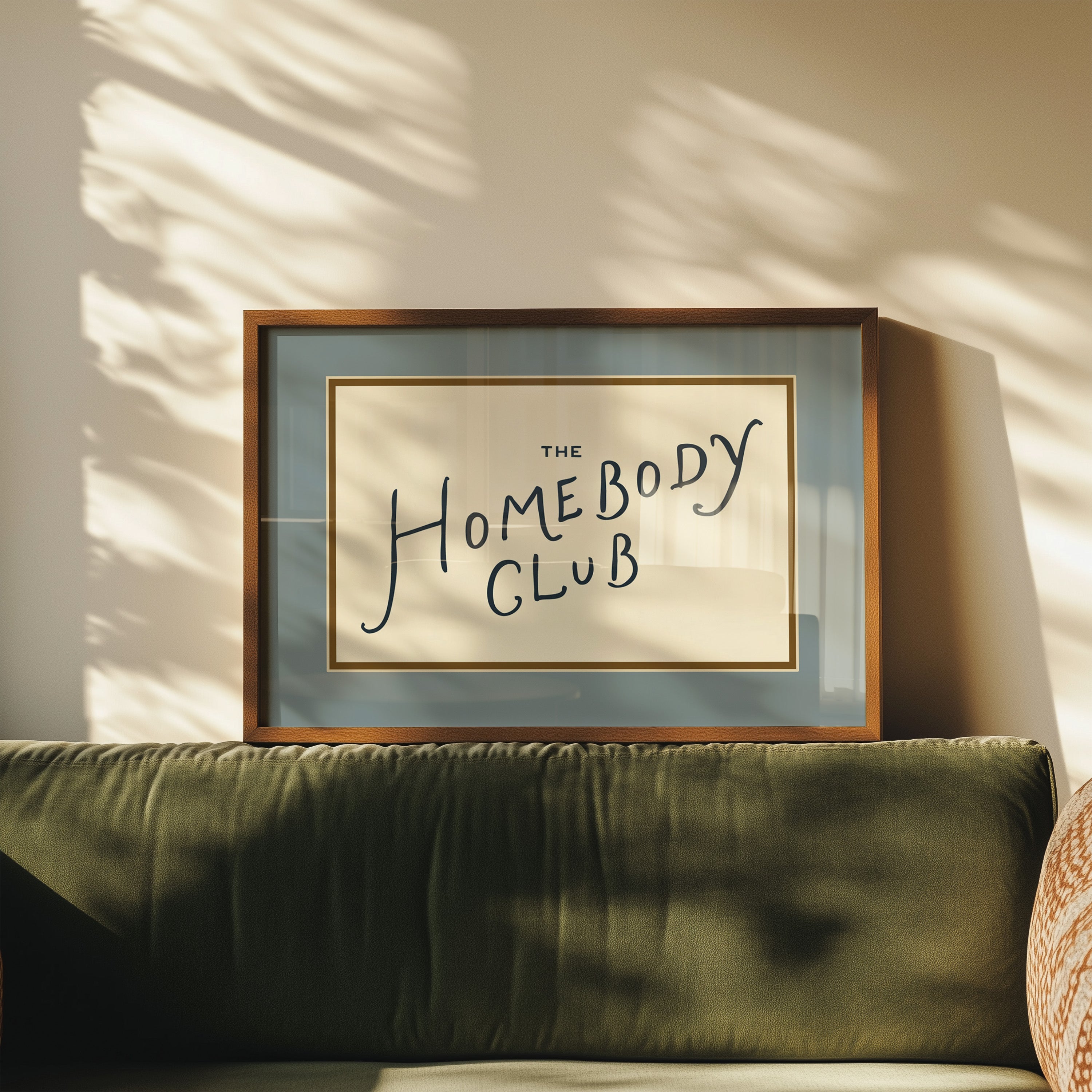 Framed artwork with 'The Homebody Club' text on a wall with sunlight casting shadows.