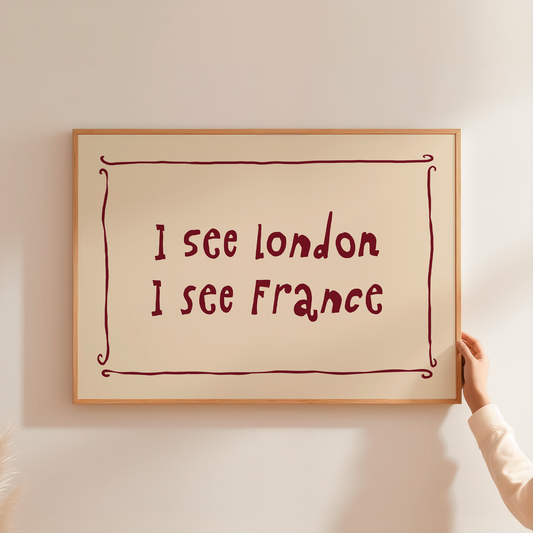 A framed typographic wall art print hanging on a light beige wall, with a hand holding the right side of the frame. The print has a cream background, a thin brown border, and dark red text reading 'I see London I see France'.