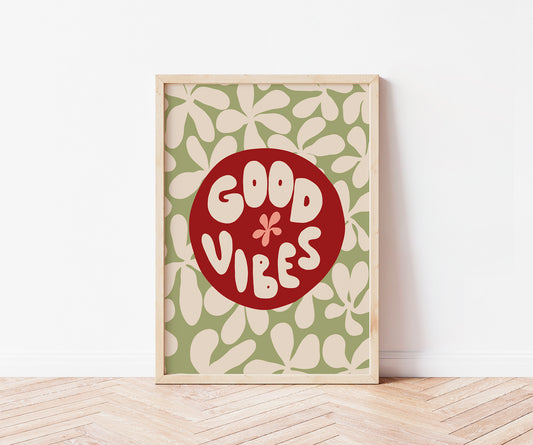 A framed art print leaning against a white wall on a light wood herringbone floor. The print features a retro design with a red circle in the center containing the words 'GOOD VIBES' in cream lettering, surrounded by a green background with cream leaf patterns.