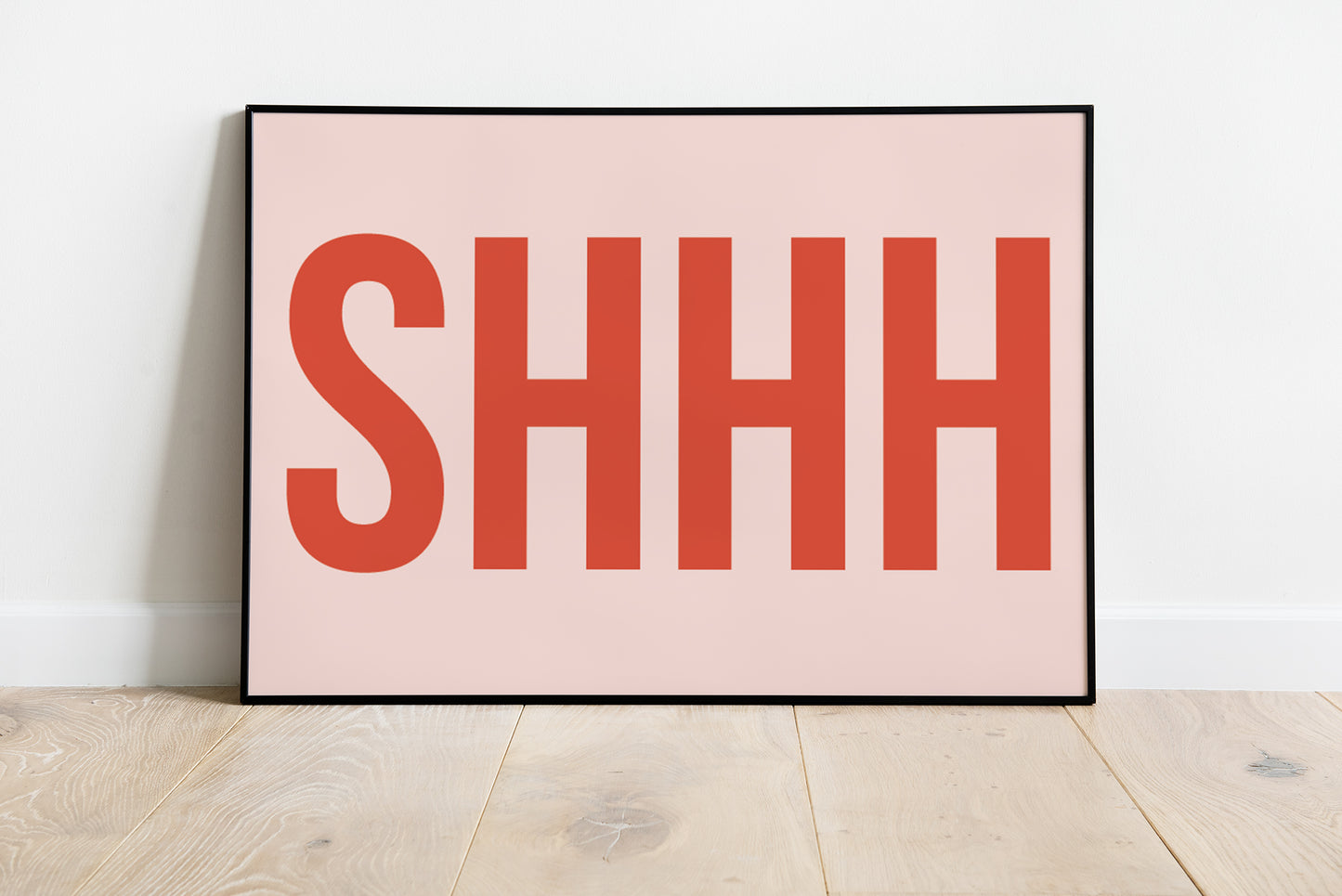 A minimalist framed art print featuring the word 'SHHH' in bold coral-red letters on a pale pink background, leaning against a white wall on light wood flooring.