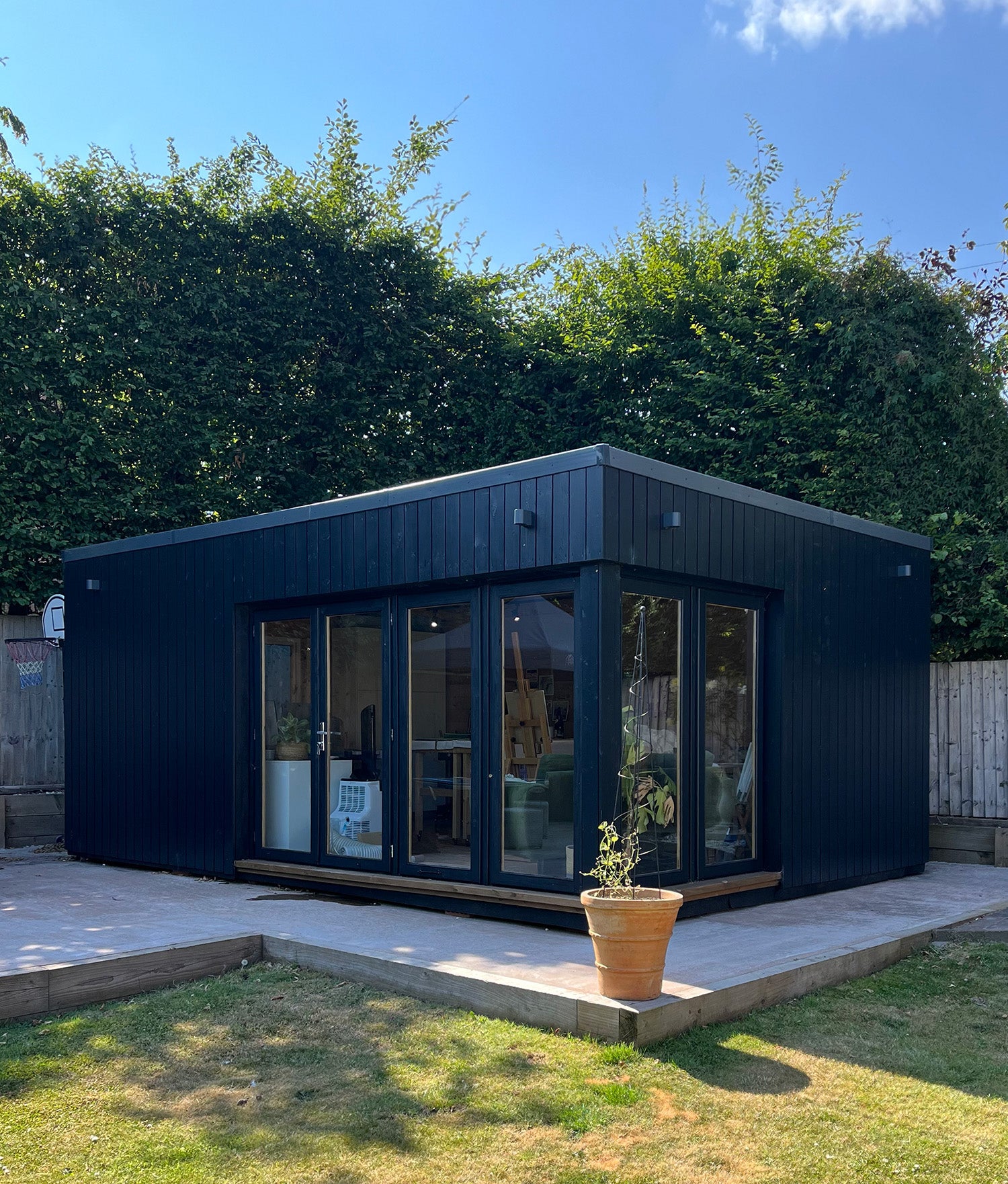 A modern black outdoor office cabin with large glass windows and doors, situated on a concrete patio in a backyard with green hedges, grass, and a blue sky.