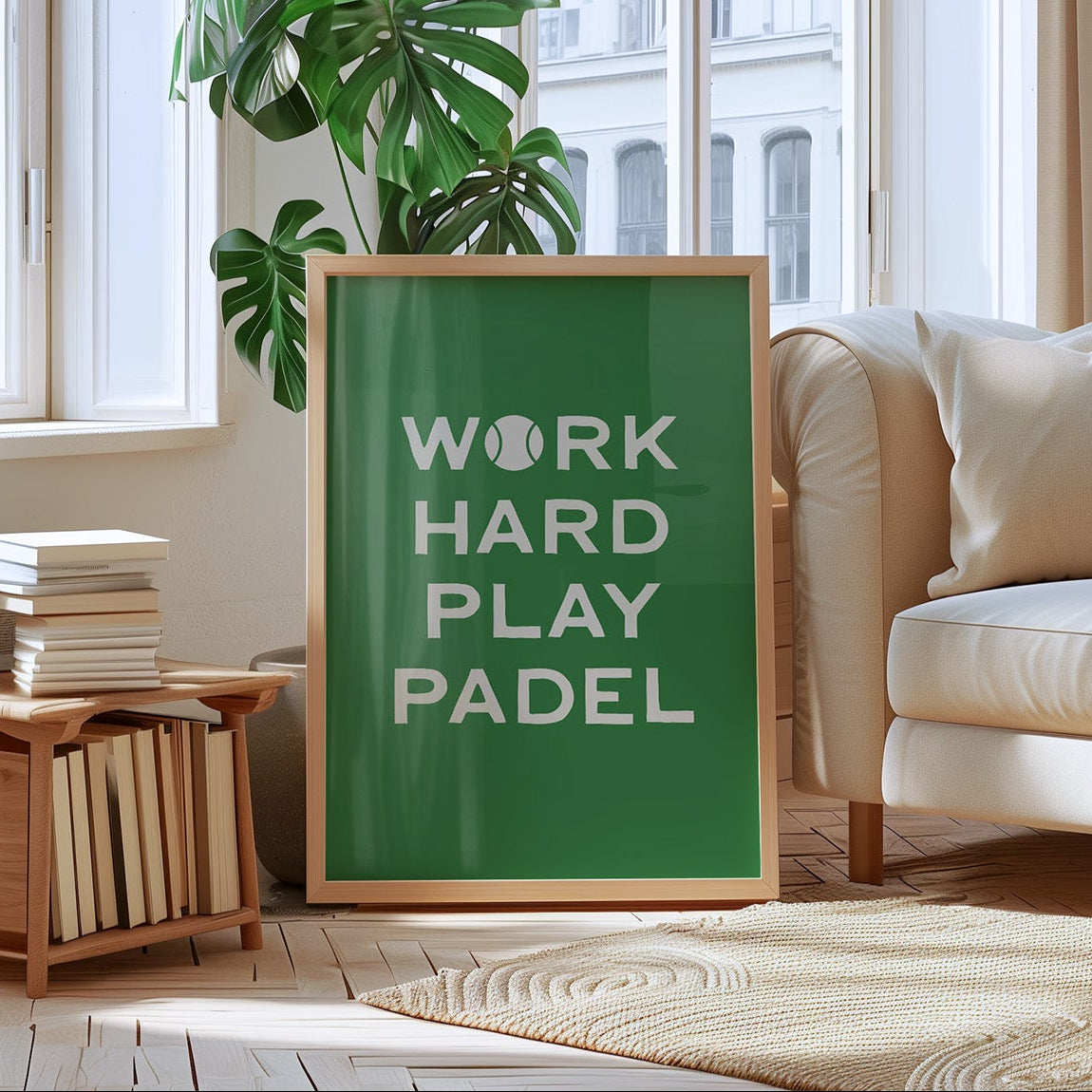 A framed green wall art print leaning in a bright living room. The print reads “WORK HARD PLAY PADEL” in white text, with a tennis ball icon replacing the O in WORK HARD.