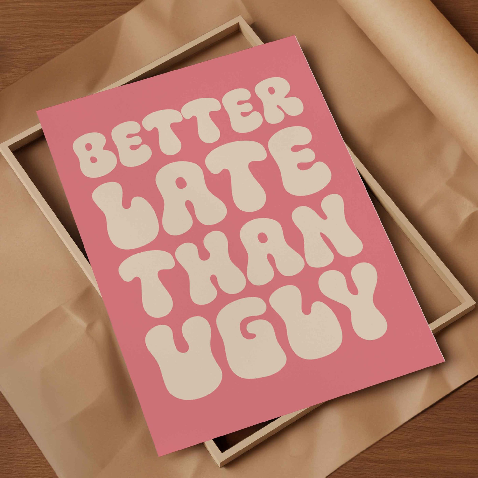 A pink art print with retro bubbly text reading 'BETTER LATE THAN UGLY' in cream, styled on top of a light wood box with brown paper inside.