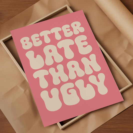 A pink art print with retro bubbly text reading 'BETTER LATE THAN UGLY' in cream, styled on top of a light wood box with brown paper inside.
